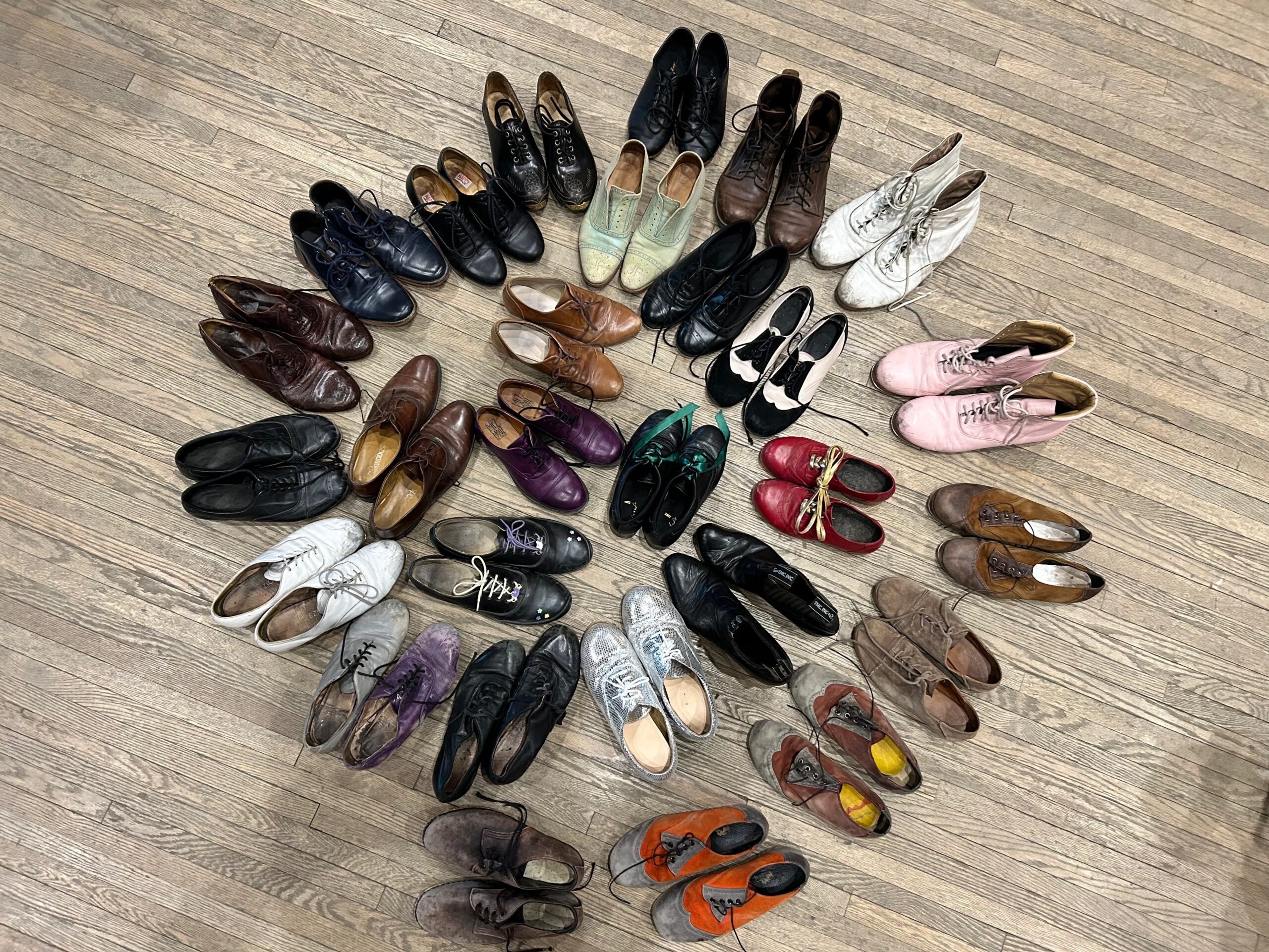 A group of hard-soled dance shoes in a variety of colors, all arranged in a rough circle on a wooden floor.