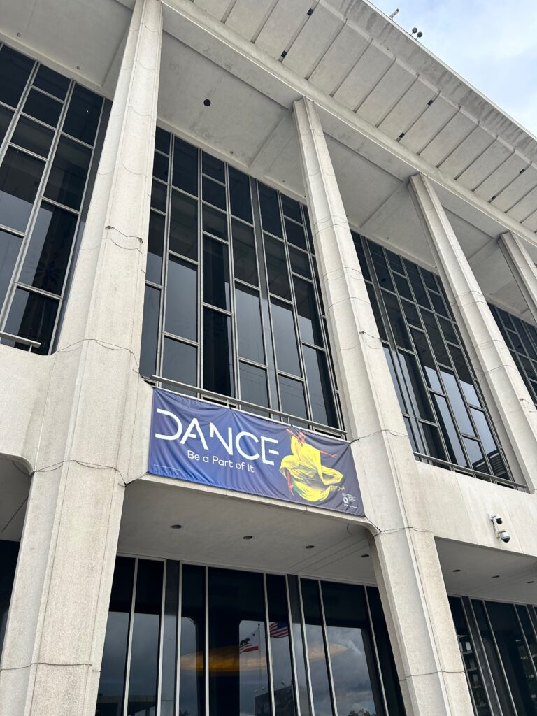 A gray stone building with tall columns and tall many-paned windows. A banner hangs over the front reading "DANCE Be a Part of It" with an image of a leaping dancer clothed in a yellow dress.