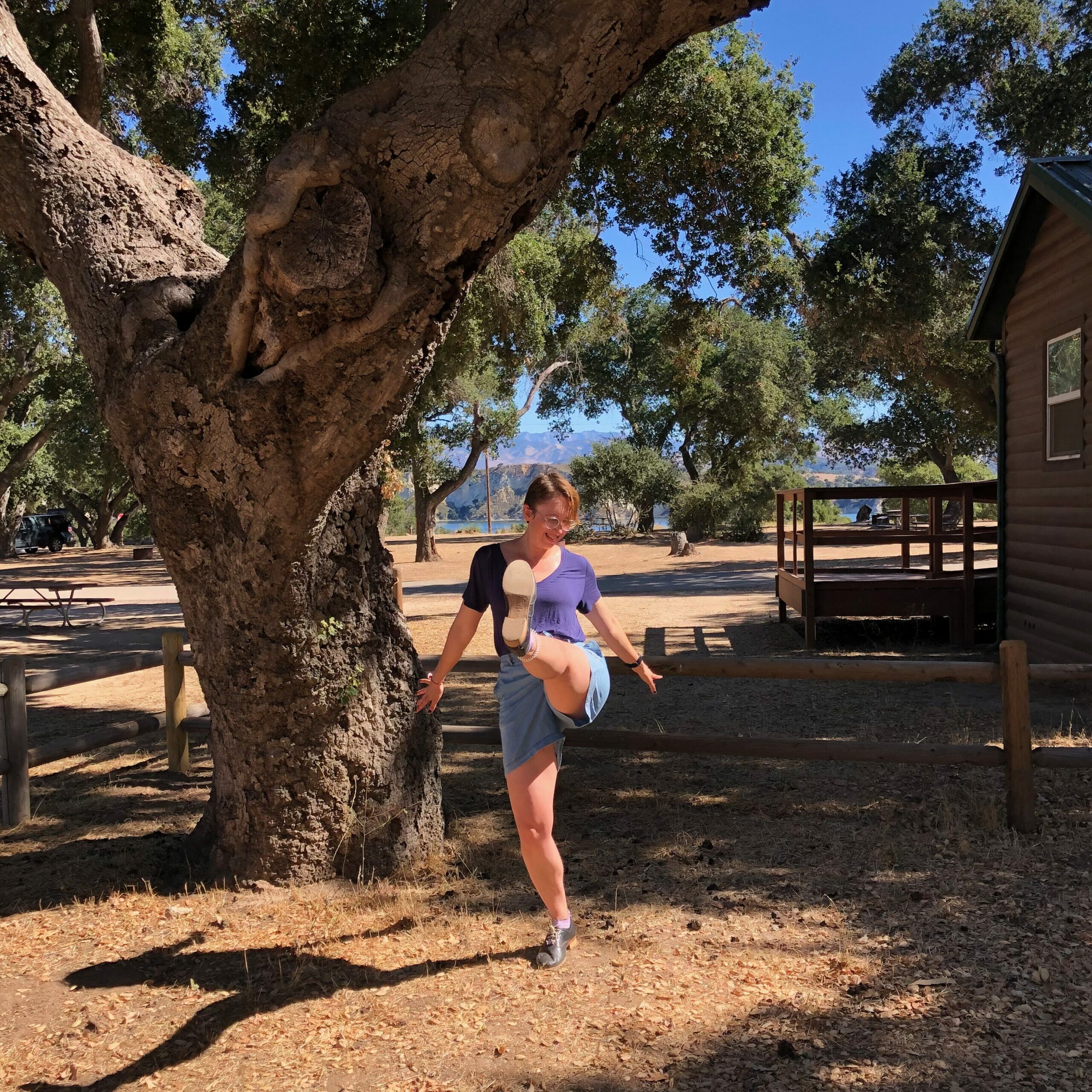 A person (me) at center, kicking one leg forward and throwing both arms back. I am standing in front of a large tree next to a wooden cabin. There is dirt stretching backwards and several trees in the distance.