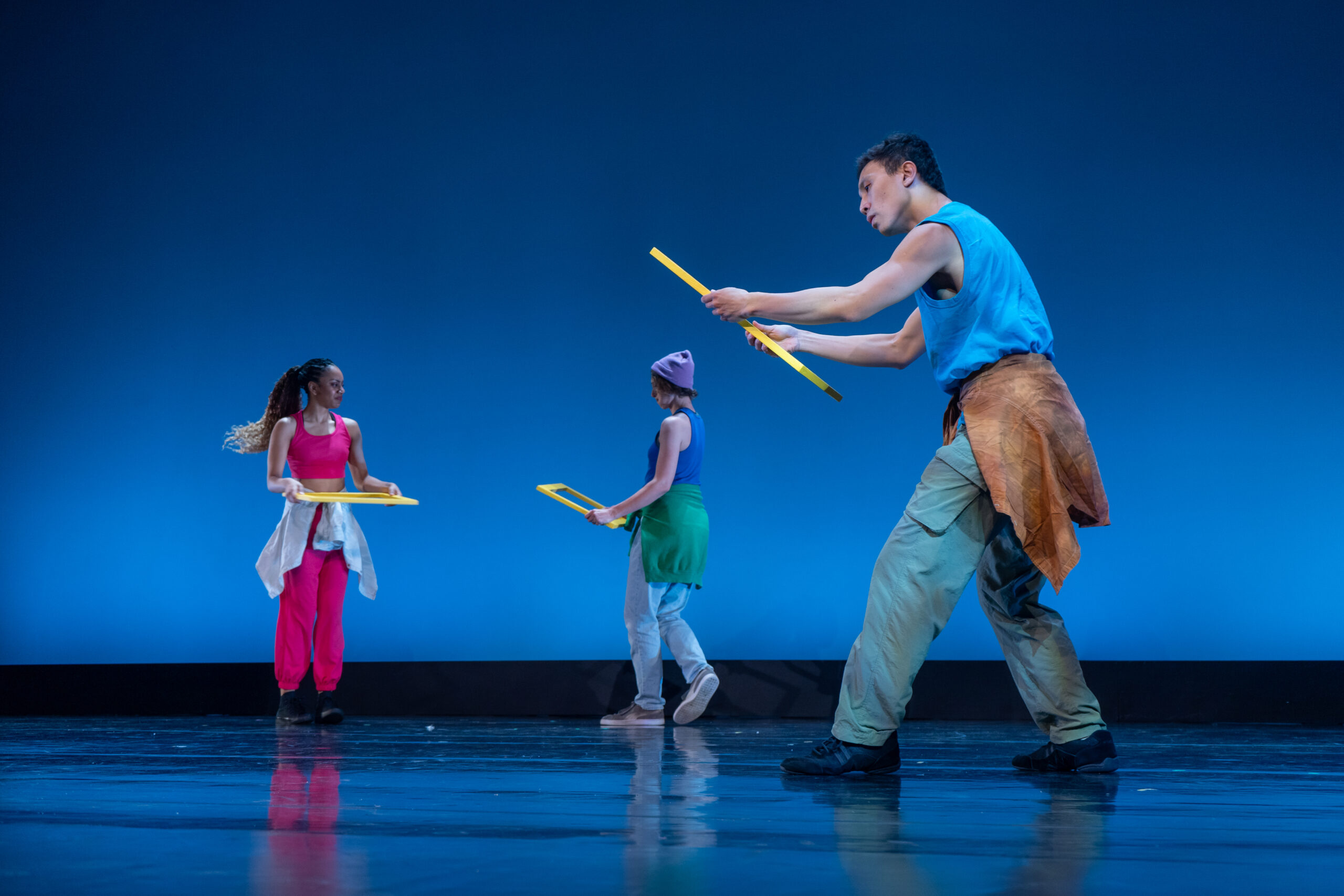 A blue-lit stage with three colorfully and casually dressed dancers. The dancers are holding yellow rectangular frames with expressions of confusion.
