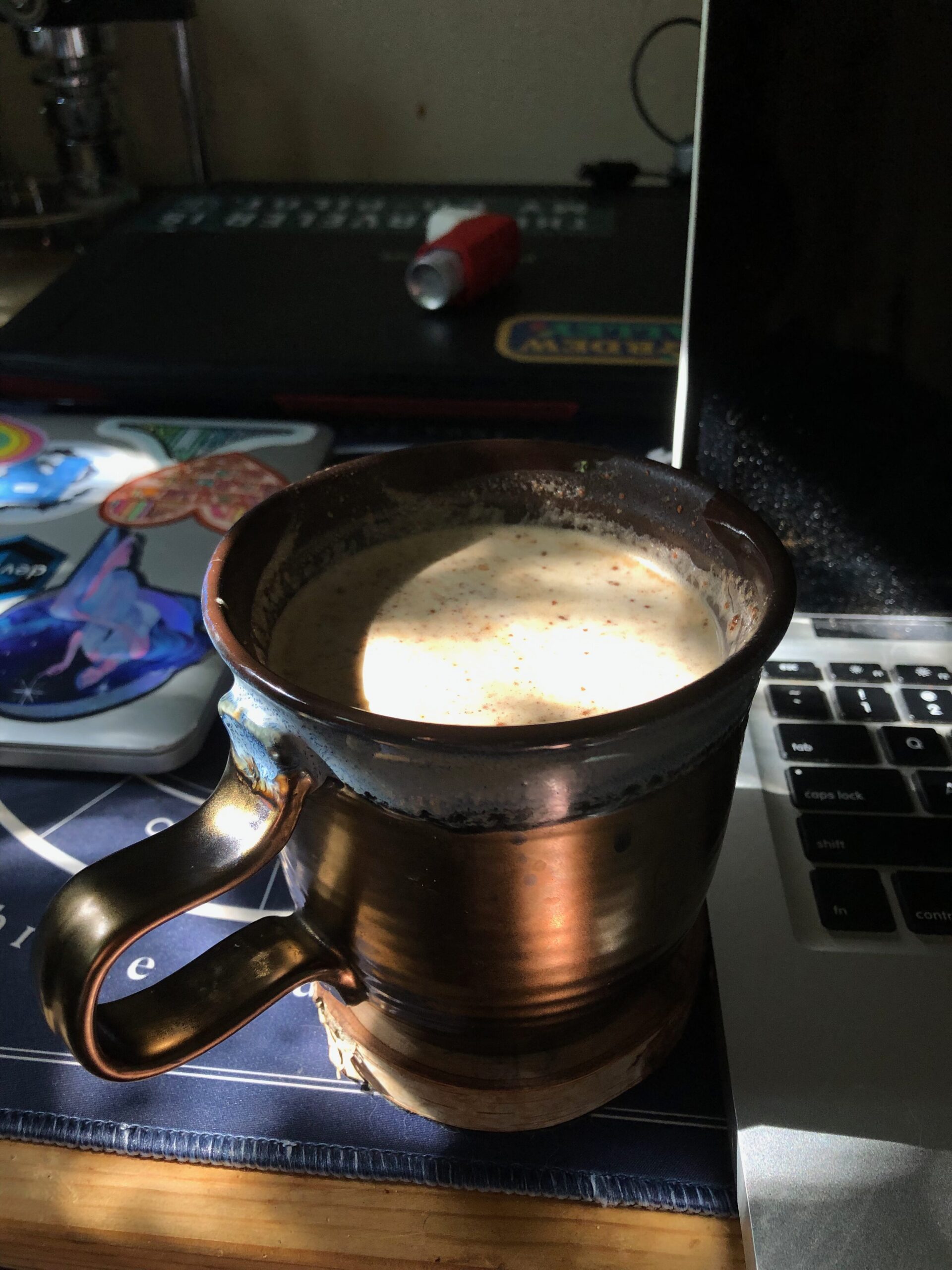A brown mug filled with a light brown, foamy latte sits next to a silver macbook laptop and on top of a blue mat printed with celestial symbols.