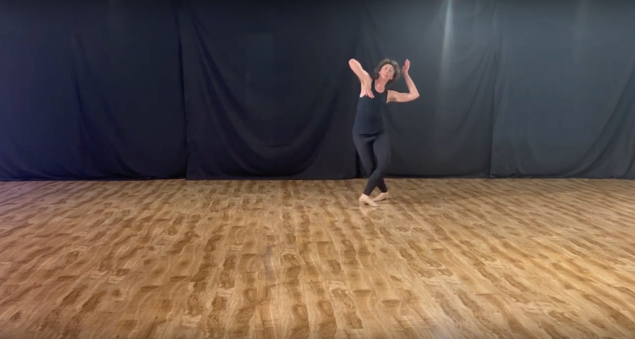 A black background, a wooden floor, and a single dancer (me) dressed in black pants and sleeveless top, leaning to the right and framing the face with one hand behind the head and one hand lifting under the chin.