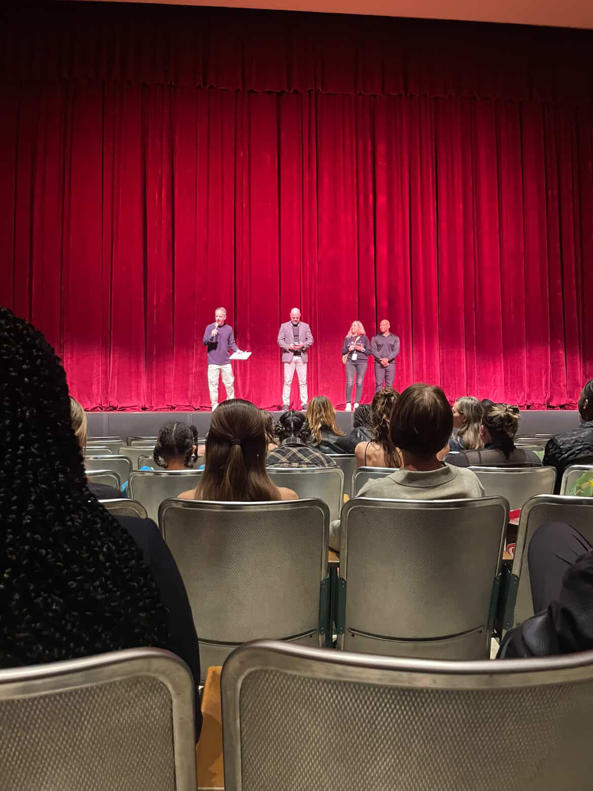 Rows of seats and the backs of audience members' heads in the foreground, with a stage and red curtain in the background. On the stage, four figures stand with one holding a microphone and speaking.